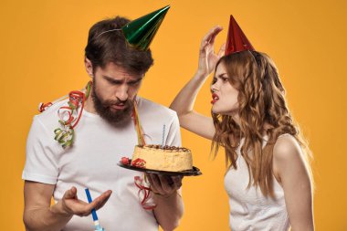 Sarı arka planda doğum gününü kutlayan şapkalı mutlu adam ve kadın ellerinde pastayla