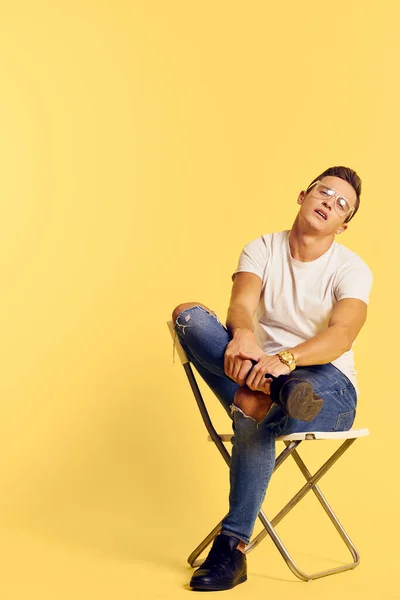 Lindo hombre sentado en una silla blanca camiseta jeans estilo moderno estilo amarillo fondo — Foto de Stock