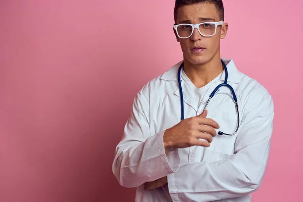 Male doctor in a medical gown with a stethoscope around his neck on a pink background and glasses on his face — Stock Photo, Image