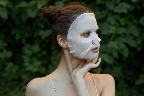 Mooi meisje cosmetische masker houdt zijn hand in de buurt van zijn gezicht en kijkt weg blote schouders bladeren op de achtergrond — Stockfoto