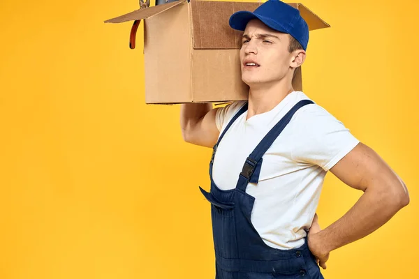 Pracujący człowiek w mundurze z pudełkiem dostawy usługi załadunku żółty tło — Zdjęcie stockowe