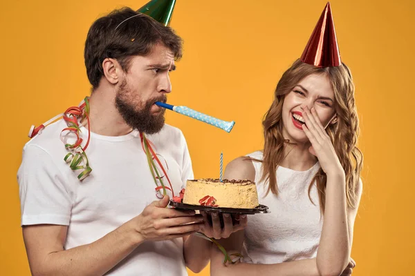 Homem e mulher felizes em um boné celebrando um aniversário em um fundo amarelo com um bolo em suas mãos — Fotografia de Stock