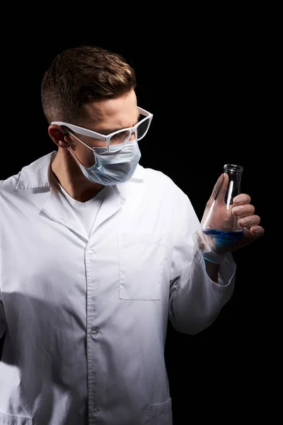 Laboratory assistant in a medical mask and in a white coat with a liquid in a flask and a chemical on a black background — Stock Photo, Image