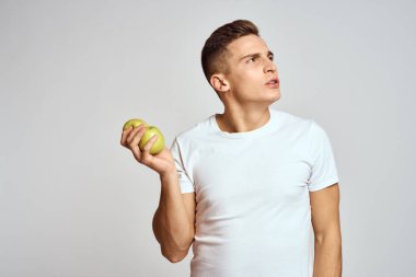 Elma tutan beyaz tişörtlü, arka planda hafif bir adam. El hareketleriyle gülümsüyor.