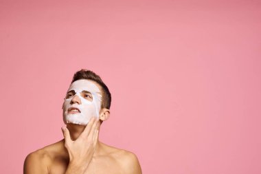 Arındırıcı maskeli bir adam yüzünde siyah noktalar, pembe arka planda kırpılmış bir manzara ve yüze yakın eller.