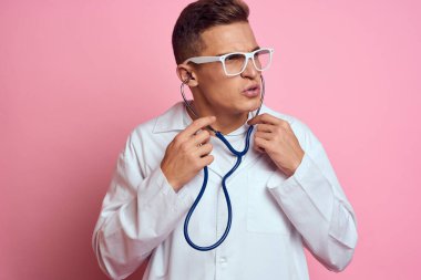 Pembe bir arka planda steteskop ve yüzünde gözlük olan tıbbi önlüklü bir erkek doktor.