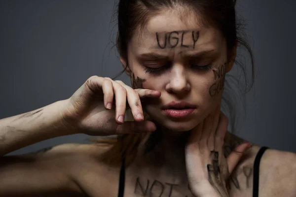 Donna frustrato con iscrizioni offensive sul suo corpo e in una t-shirt nera su un ritratto di sfondo grigio — Foto Stock
