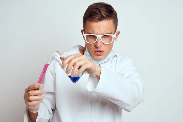 Un assistant de laboratoire en blouse médicale avec un masque de protection tient une fiole avec un liquide de réaction chimique — Photo
