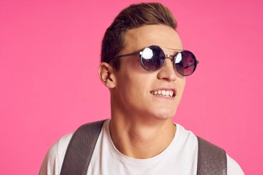 Güneş gözlüklü, beyaz tişörtlü, pembe arka planlı bir adam.