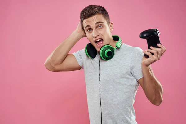 Ένας άνδρας με ένα χειριστήριο στα χέρια του ακουστικά που παίζουν παιχνίδια ψυχαγωγία lifestyle λευκό t-shirt ροζ φόντο — Φωτογραφία Αρχείου