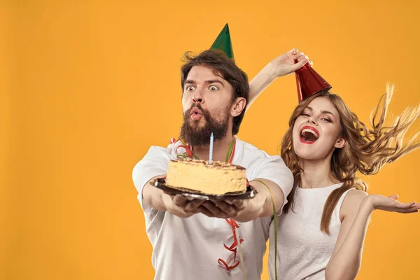 Glad man och kvinna i en mössa firar en födelsedag på en gul bakgrund med en kaka i händerna — Stockfoto
