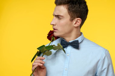 Tişörtlü ve kravatlı bir adam. Omzunda kırmızı bir gül, sarı bir arka planda.