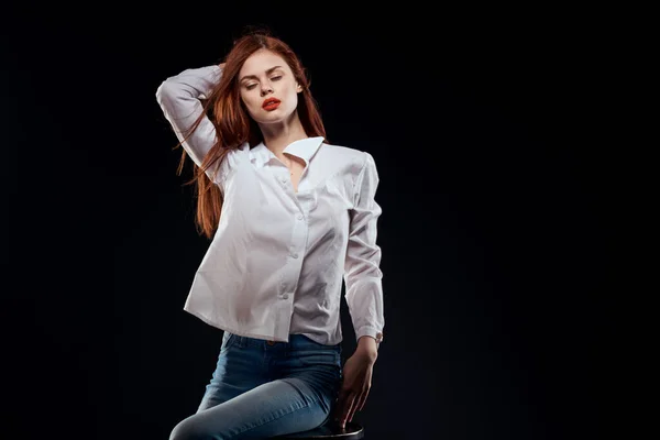 Pretty woman sitting on a chair posing white shirt jeans long hair red lips dark background — Stock Photo, Image