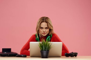 Kulaklık takan, dizüstü bilgisayarlı bir kadın masa başı eğlence sektöründe pembe arka planda oturuyor.