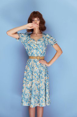 Çiçek elbiseli bir kadın ileriye bakıyor. Yüzüne yakın bir el tutuyor. 