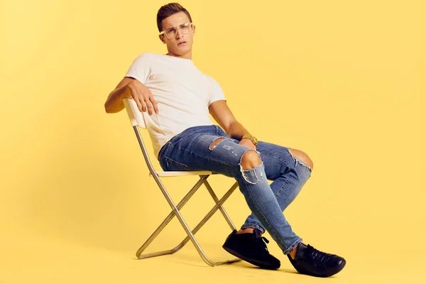 Lindo hombre sentado en una silla blanca camiseta jeans estilo moderno estilo amarillo fondo — Foto de Stock