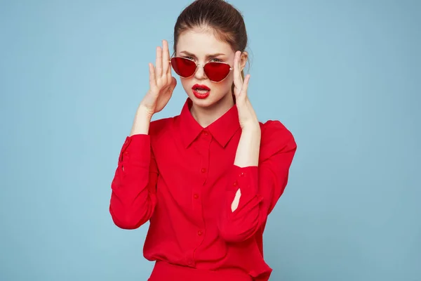 Mujer con camisa roja sobre fondo azul sostiene las manos cerca de la cara y gafas de sol labios rojos modelo de maquillaje — Foto de Stock