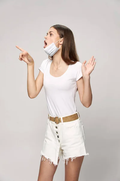 Meglepett nő orvosi maszkkal az arcán rövidnadrágban és egy pólóban mutatja az ujját. — Stock Fotó