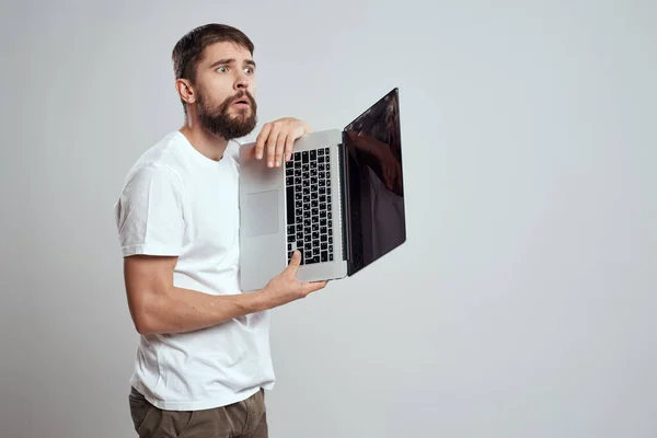 Um homem com um laptop em suas mãos em um fundo leve em uma camiseta branca emoções luz de fundo novas tecnologias — Fotografia de Stock