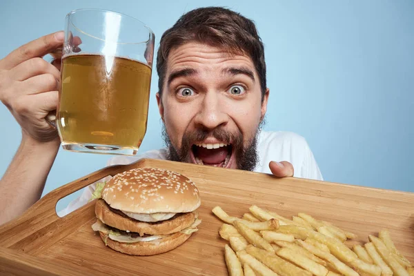 Homem alegre caneca de cerveja hambúrguer batatas fritas fast food dieta azul fundo close-up — Fotografia de Stock