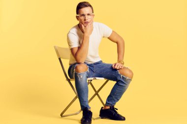 Sandalyede oturan yakışıklı adam beyaz tişört kot pantolon modern tarz sarı arka plan