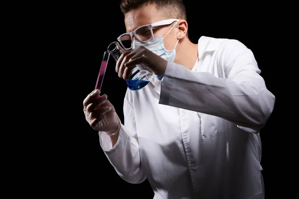 Asystent laboratoryjny w masce medycznej i w białej powłoce z płynem w kolbie i substancją chemiczną na czarnym tle — Zdjęcie stockowe