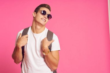 Güneş gözlüklü, beyaz tişörtlü, pembe arka planlı bir adam.