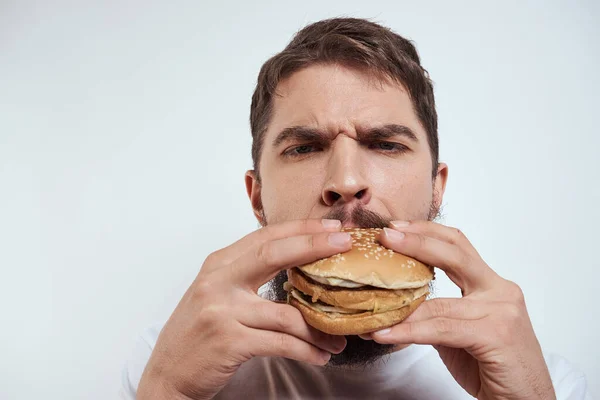 Beyaz bir tişörtle arka planda hamburger yiyen bir adam yakın çekim açlık ve fast food manzarası izledi. — Stok fotoğraf