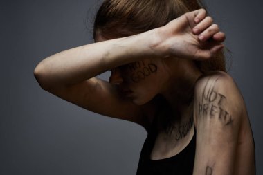 Hüsrana uğramış bir kadın vücudunda çirkin yazıtlar ve gri bir arka plan portresi üzerinde siyah bir tişört var. 