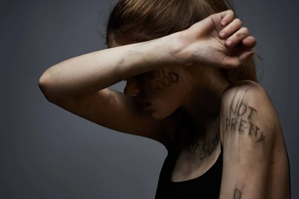 Mulher frustrada com inscrições ofensivas em seu corpo e em uma camiseta preta em um retrato de fundo cinza — Fotografia de Stock