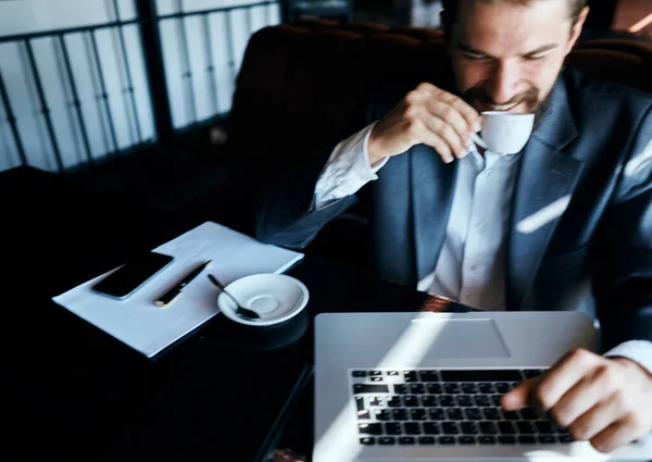 Homem de negócios sentado em um café com uma xícara de café em suas mãos na frente de uma comunicação de tecnologia de laptop — Fotografia de Stock