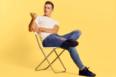 Sandalyede oturan yakışıklı adam beyaz tişört kot pantolon modern tarz sarı arka plan