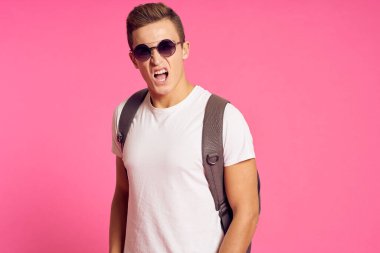 Beyaz tişörtlü ve sırt çantasında güneş gözlüğü olan pembe arka plan modeli olan bir adamın portresi.