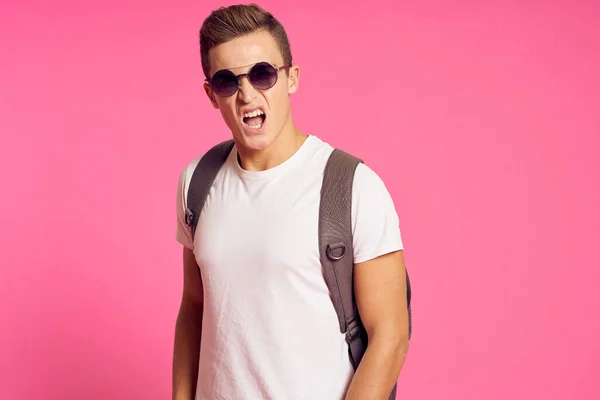 Retrato de um homem em uma camiseta branca e com uma mochila em seus óculos de sol traseiros modelo de fundo rosa — Fotografia de Stock