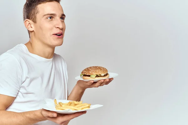 Ein Mann mit Pommes und einem Hamburger auf hellem Hintergrund in weißem T-Shirt in Großaufnahme — Stockfoto
