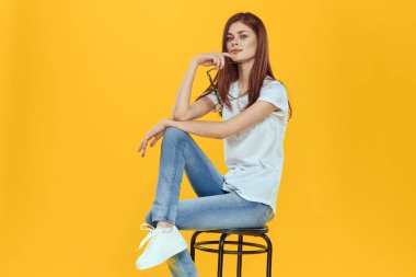 Bir kadın sandalyeye oturur ve elinde siyah bardaklarla beyaz tişörtlü kadınların moda kıyafetleri sarı arka planda kalır.