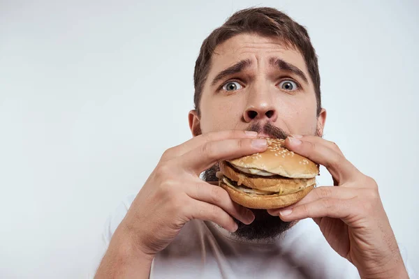 Ein Mann, der einen Hamburger auf hellem Hintergrund in einem weißen T-Shirt isst, hat die Nahaufnahme von Hunger-Fast Food abgeschnitten — Stockfoto