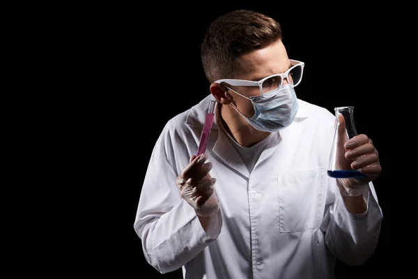 Laboratorieassistent i medicinsk mask och i vit rock med vätska i en kolv och en kemikalie på svart bakgrund — Stockfoto
