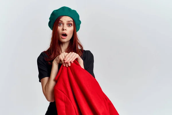 Bir kadın elinde kırmızı bir palto ve koyu renk bir tişört, açık arkaplan manzaralı yeşil bir şapka tutuyor. — Stok fotoğraf