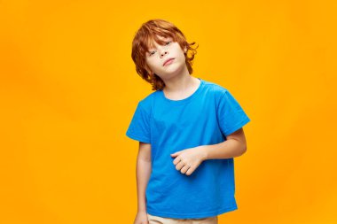 Kızıl saçlı çocuk elini mavi bir tişörtün üzerinde tutuyor. Başını bir tarafa eğerek sarı izole bir arka plan oluşturuyor. 