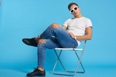 Kotlu, dizleri yırtık rahat bir adam mavi arka planda bir sandalyede oturuyor ve yüzünde uzun gözlüklerle oturuyor.