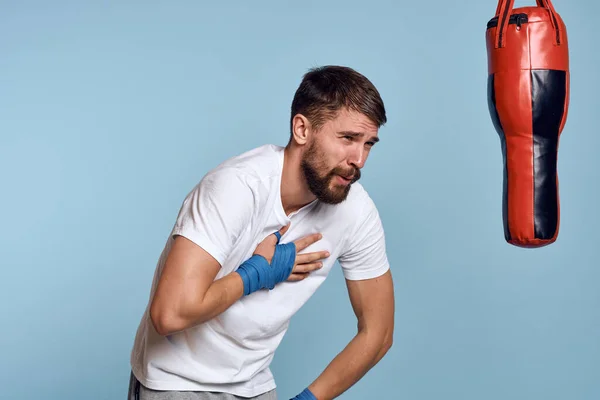 Un homme pratiquant un coup de poing sur un sac de boxe dans un t-shirt blanc sur un fond bleu — Photo