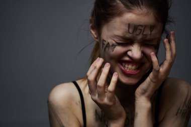 Gri arka planda kırıcı yazılar yazan üzgün kadın elleriyle kendine dokunuyor.