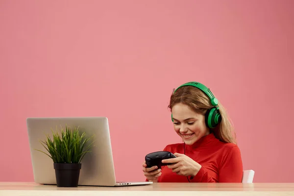 Mulher em fones de ouvido com gamepad na frente do laptop sentar na mesa de entretenimento estilo de vida rosa fundo — Fotografia de Stock