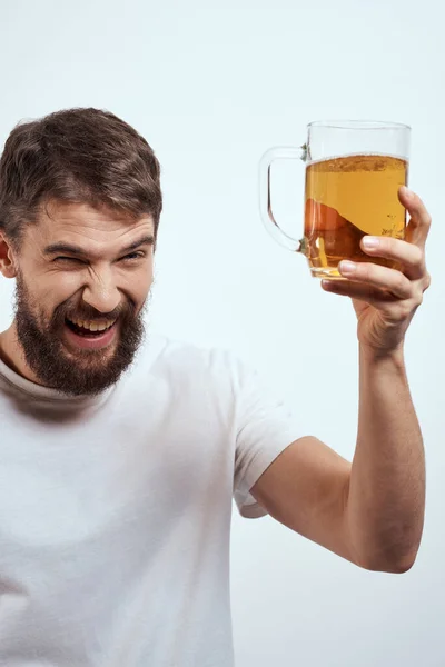 Un homme barbu avec une tasse de bière sur un fond clair dans un T-shirt blanc vue recadrée d'une boisson alcoolisée — Photo