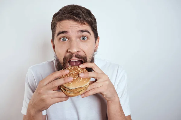 Muž jíst hamburger na světlém pozadí v bílém tričku oříznutý pohled zblízka hlad fast food — Stock fotografie