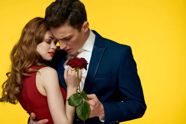 Egy romantikus férfi megölel egy vörös ruhás nőt egy rózsával a kezében, sárga alapon. — Stock Fotó