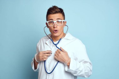Gecelik ve gözlüklü bir sağlık çalışanı mavi arka planda steteskop tutuyor.