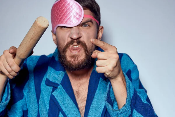 Un hombre con una máscara de sueño rosa sostiene un rodillo en sus manos y emociones energía agresión modelo bata azul — Foto de Stock