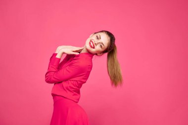 Pembe arka planda, şık elbiseli ve parlak makyajlı bir kadın.